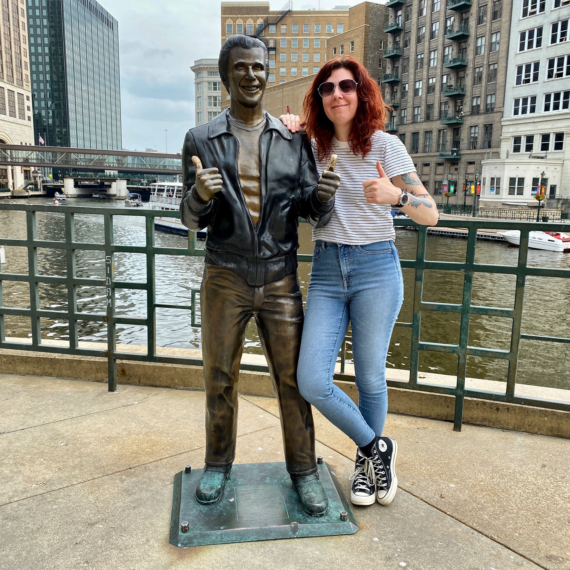 A white woman with long dark reddish-brown hair wearing blue jeans, striped t-shirt and black Converse stands next to a bronze statue of the Fonz, a man wearing a leather jacket and holding thumbs up, in front of the river in downtown Milwaukee