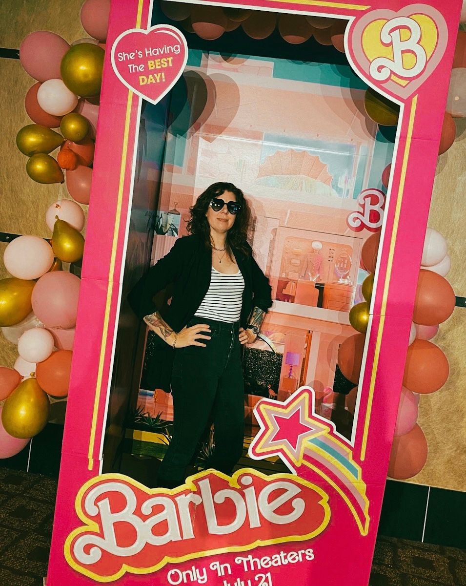A white woman in a black jacket, striped top and black pants poses with hands on hips and black heart sunglasses inside of a life-size pink Barbie doll box