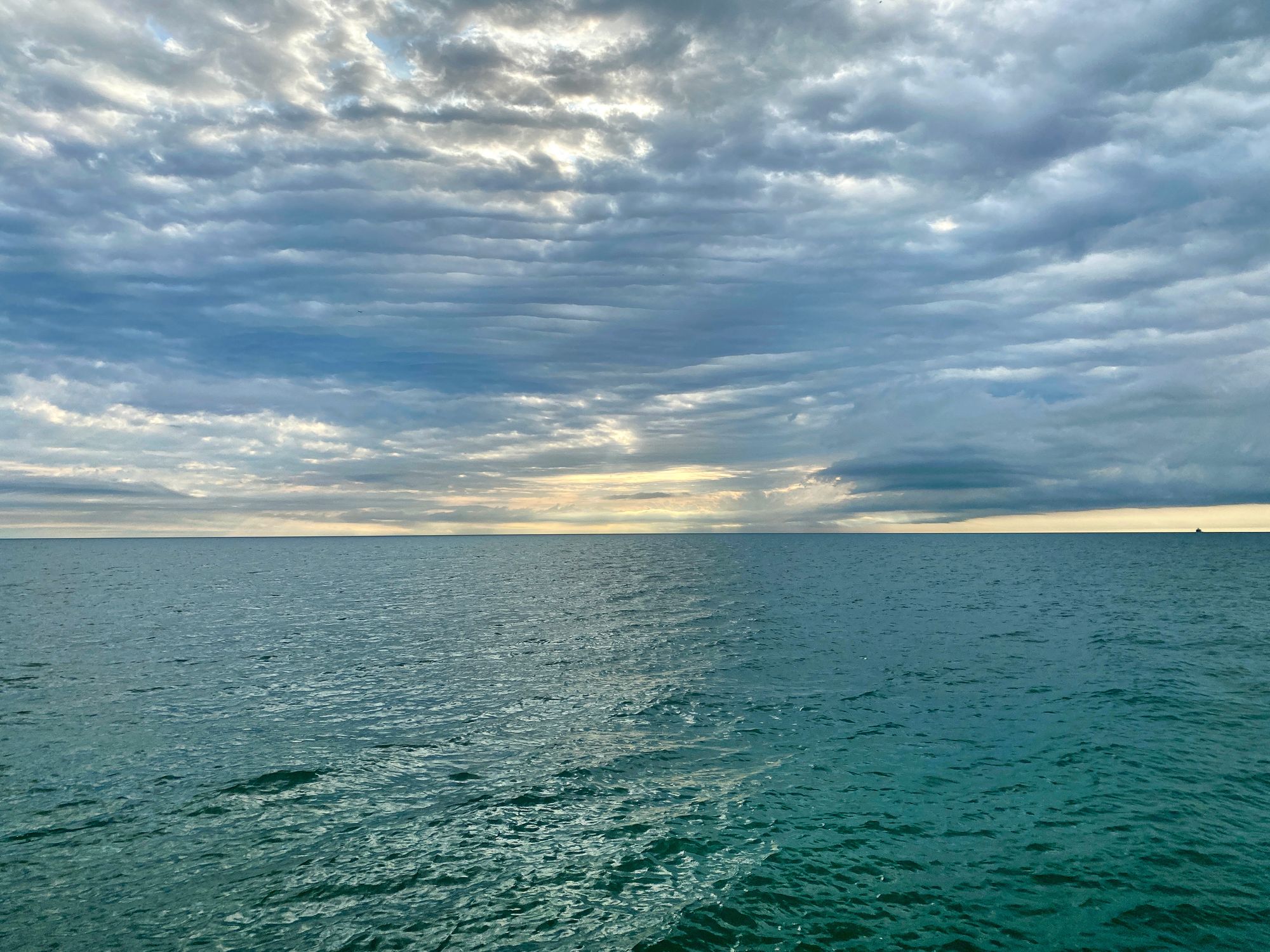 A wide horizon with ripples of clouds and wisps of yellow sunrise in the sky above a deep green lake surface