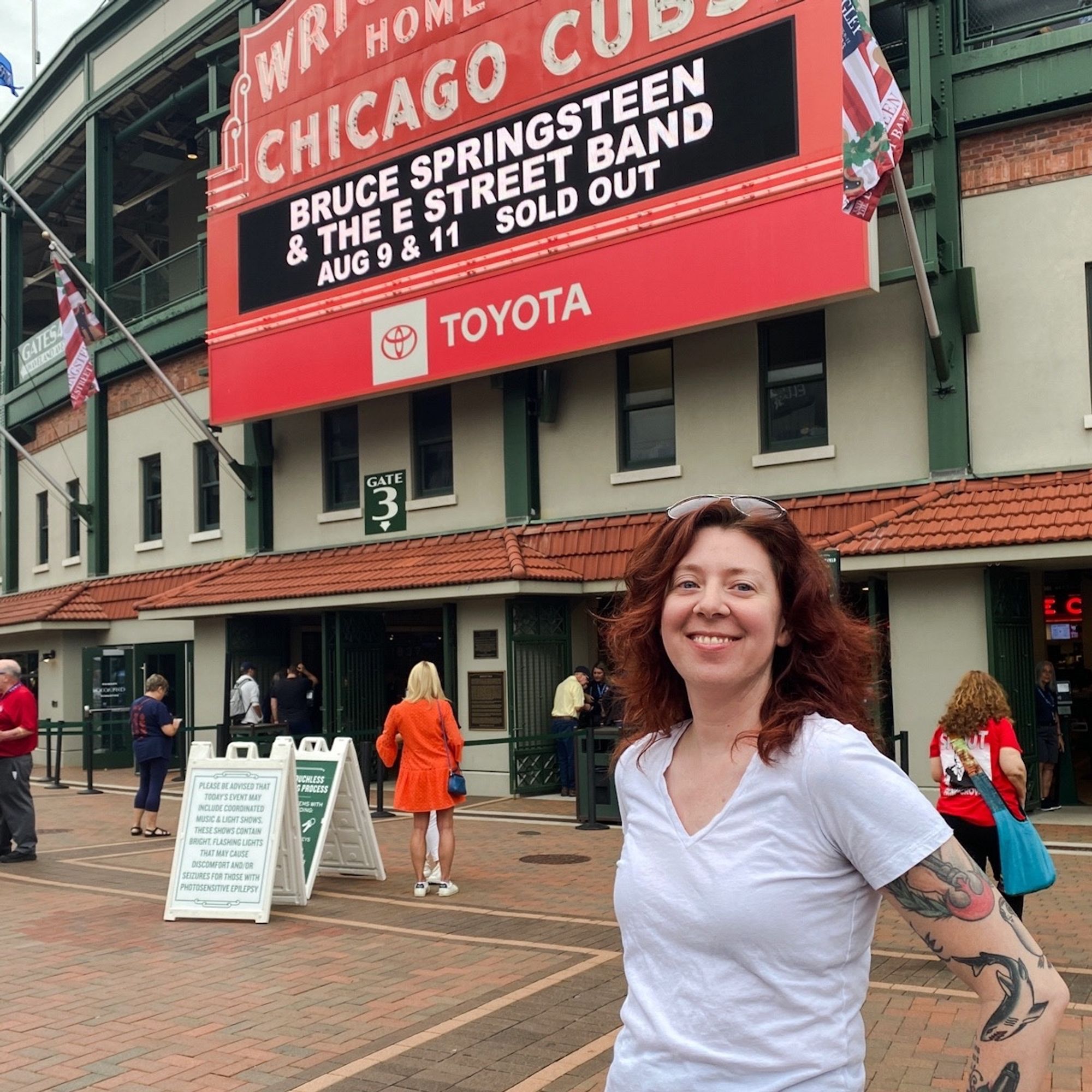 White woman with long, wavy dark red hair smiles in front of Wrigley Field stadium with sign reading “Bruce Springsteen and the E-Street Band Aug 9 & 11 Sold Out”