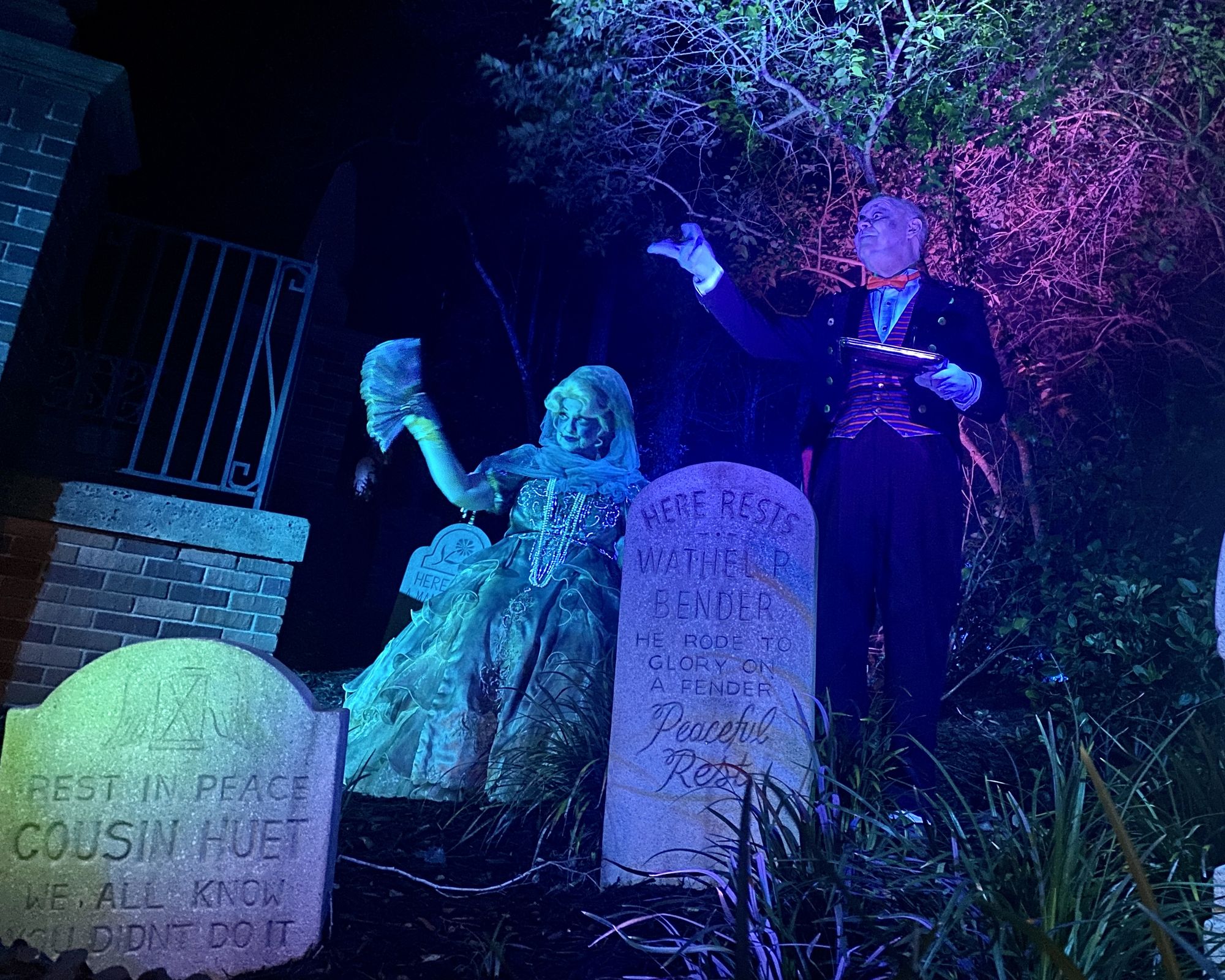 Live actors at Disney World's Haunted Mansion, dressed as a fine lady and a butler, lit in dramatic green and purple, next to tombstones