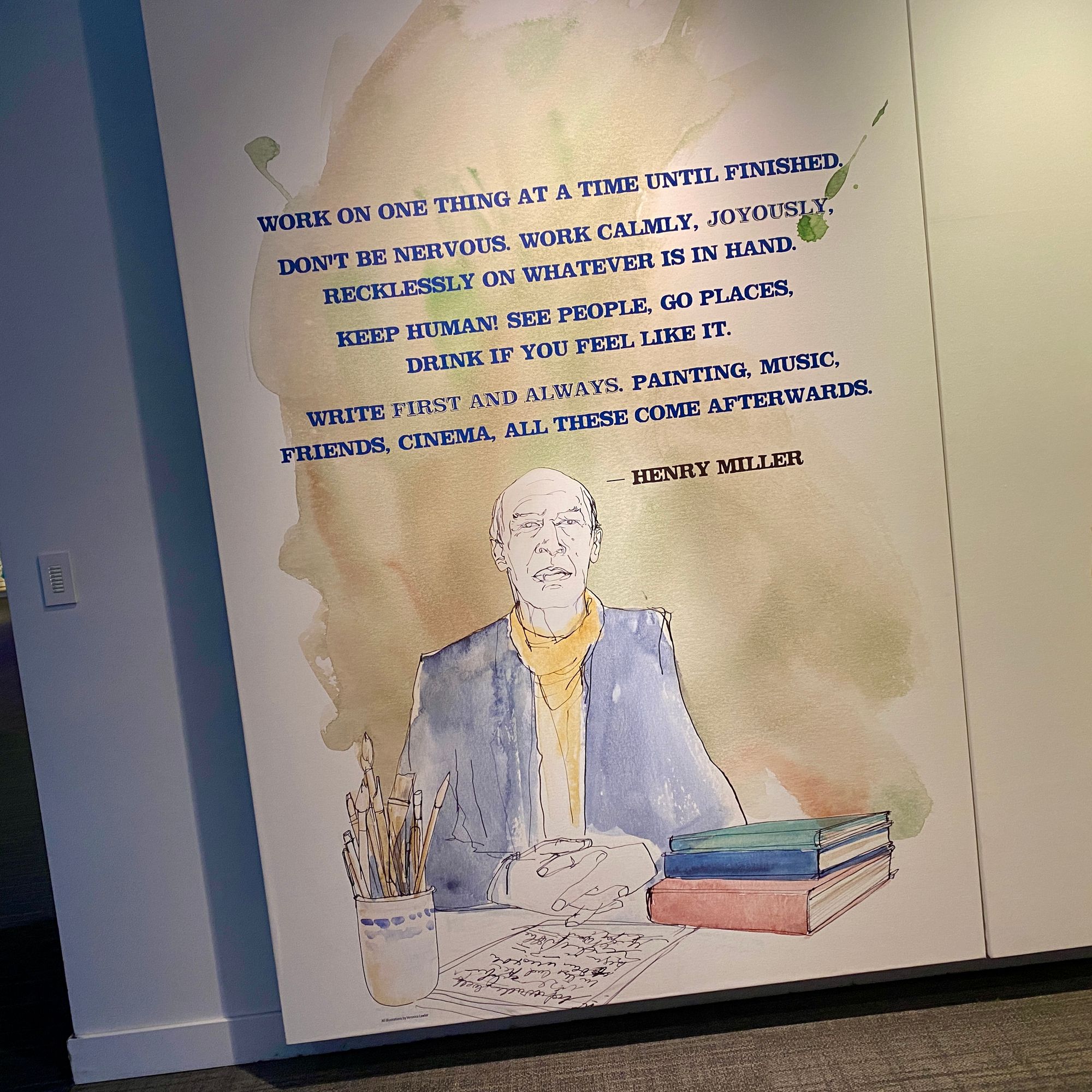 Portion of museum wall with a watercolor image of Henry Miller, an older white man, with the quote: "Work on one thing at a time until finished. Don't be nervous. Work calmly, joyously, recklessly on whatever is at hand. Keep human! See people, go places, drink if you feel like it. Write first and always. Painting, music, friends, cinema, all these come afterwards."