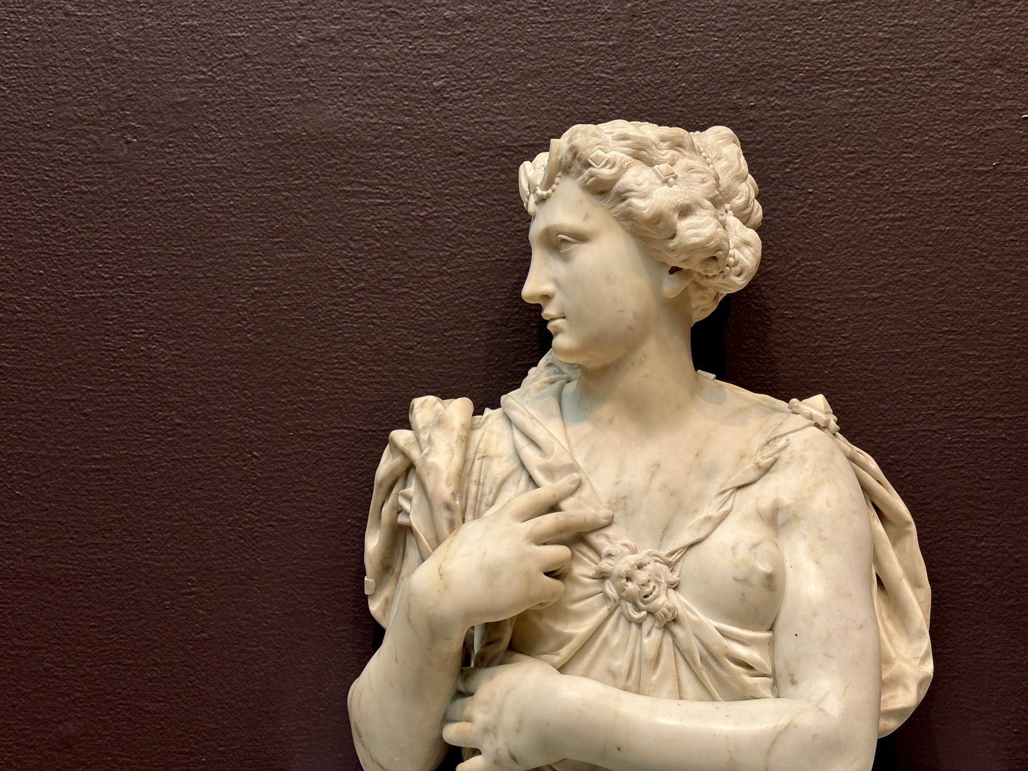 A marble bust of a woman in Grecian dress, head turned to the side and hair dressed with pearls, against a dark painted wall