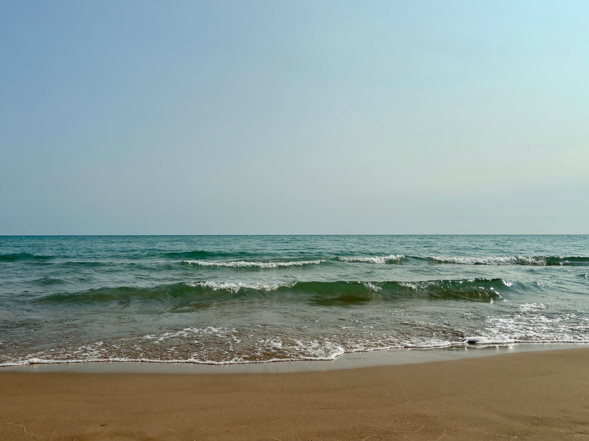 Wide lake horizon with clear pale blue sky above and below blue-green waves breaking onto a sandy beach