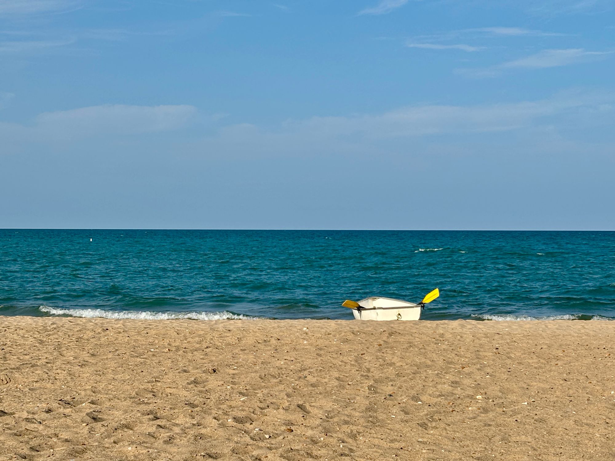 A pale blue sky swept with thin clouds over a deep turquoise lake, with waves rolling up on a sandy beach, a white rowboat with yellow oars at the edge of the water