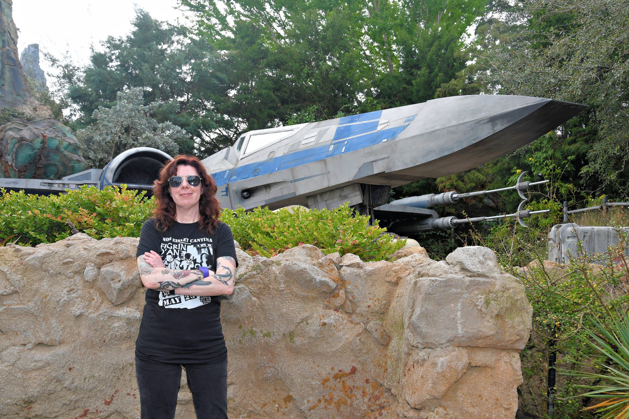 Me, a white woman with long wavy dark red hair and tattooed arms, wearing aviator sunglasses, black t-shirt and black jeans, standing in front of a X-Wing fighter from Star Wars.