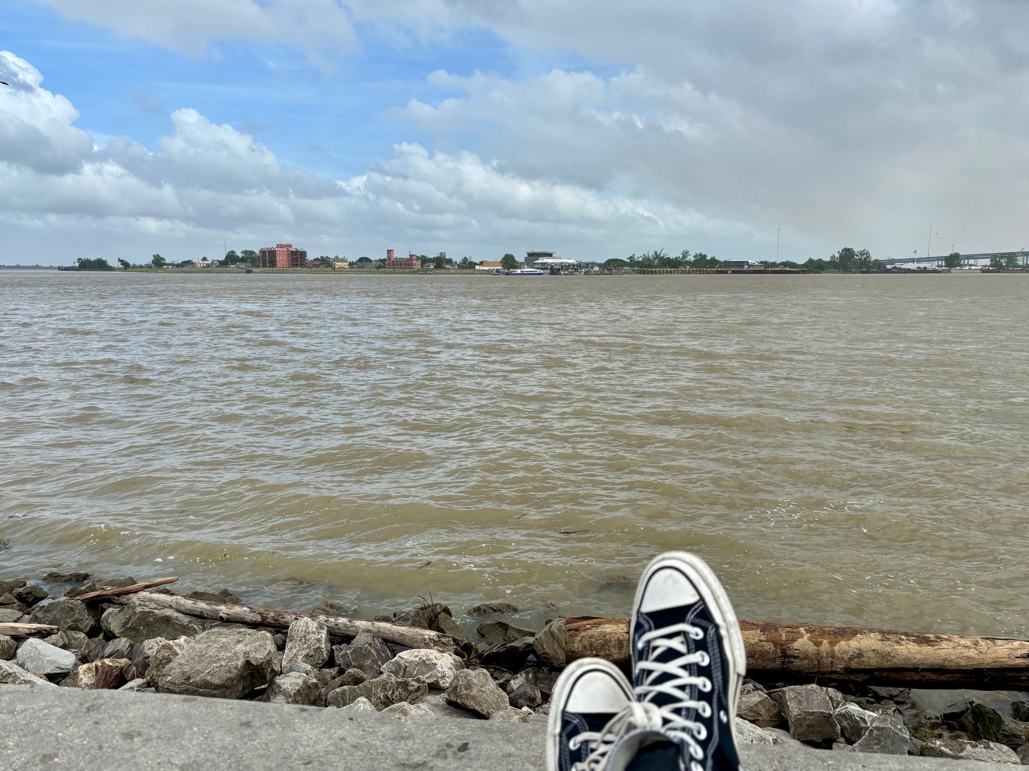 A clouded sky with bits of blue over the expanse of the muddy Mississippi River, a pair of black Converse shoes crossed at the bottom of the frame.