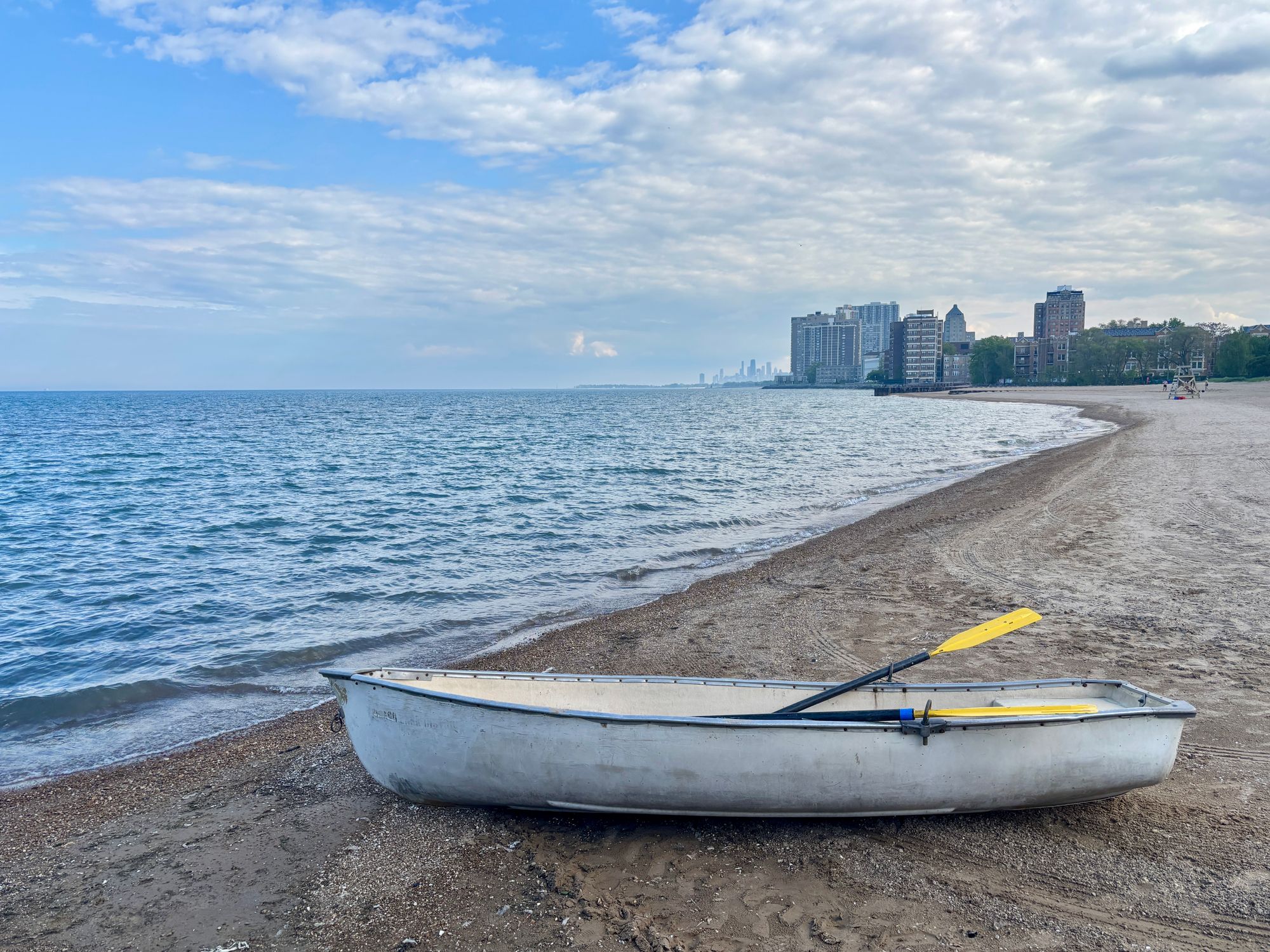 A white rowboat with yellow oars set on a sandy beach along the edge of a wide blue lake curving into the distance with the Chicago skyline in the distance under a pale blue sky swept with clouds.